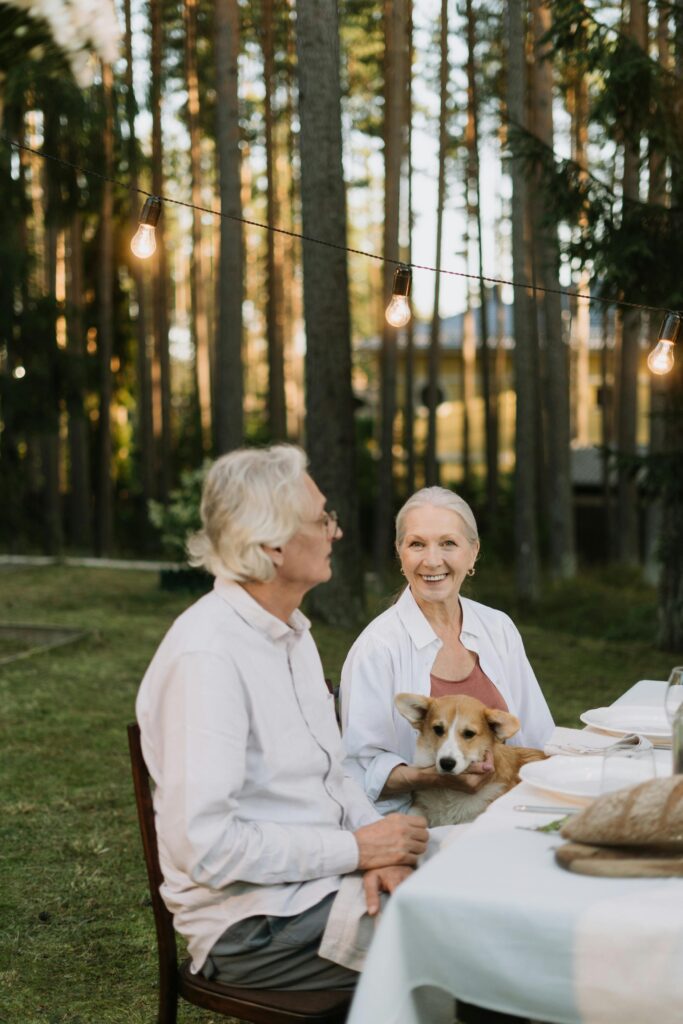 pexels-photo-5961944-5961944 Elderly couple dining outdoors with a corgi, embracing nature and happiness.